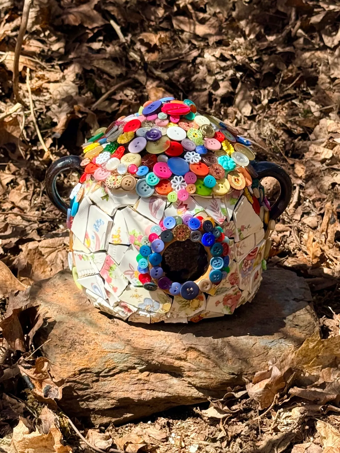 A round faerie house covered in colourful buttons, resting on the forest floor.