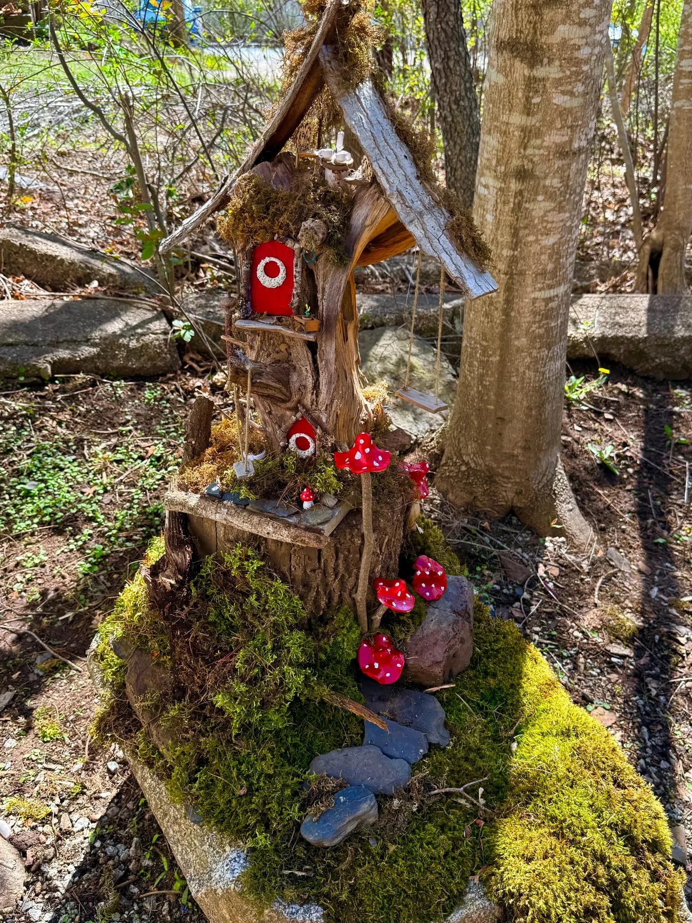 A handcrafted faerie house with a mossy bark roof and red toadstools, tucked into a tree beside a woodland stream.